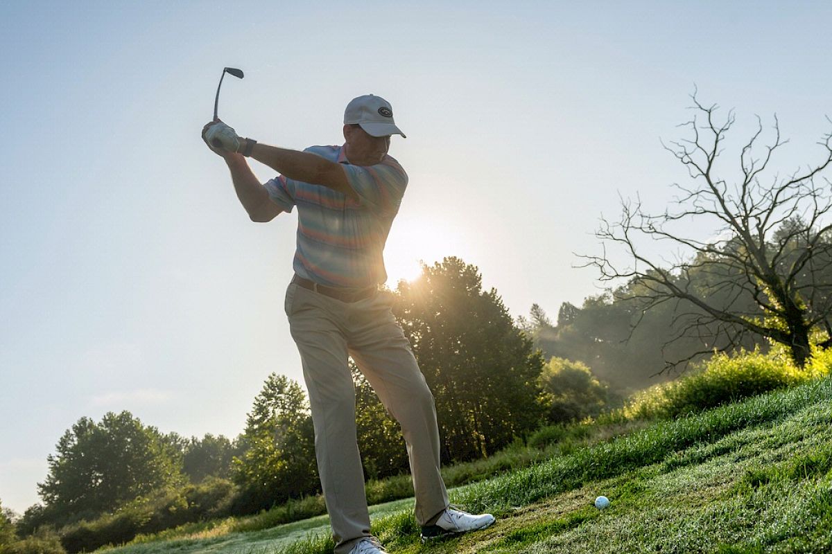 A man swings a golf club on a grassy course at sunrise, with trees and a bare tree in the background, wearing a cap and casual golf attire.