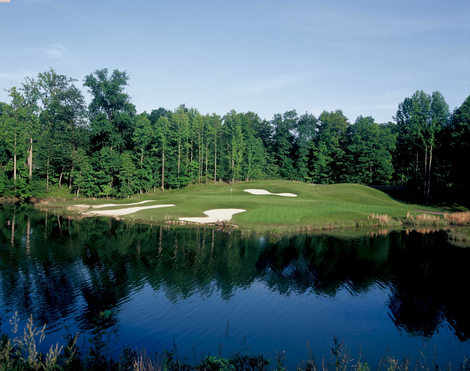 A serene golf course next to a reflecting pond, with lush greenery and sand traps in the background, under a clear blue sky.