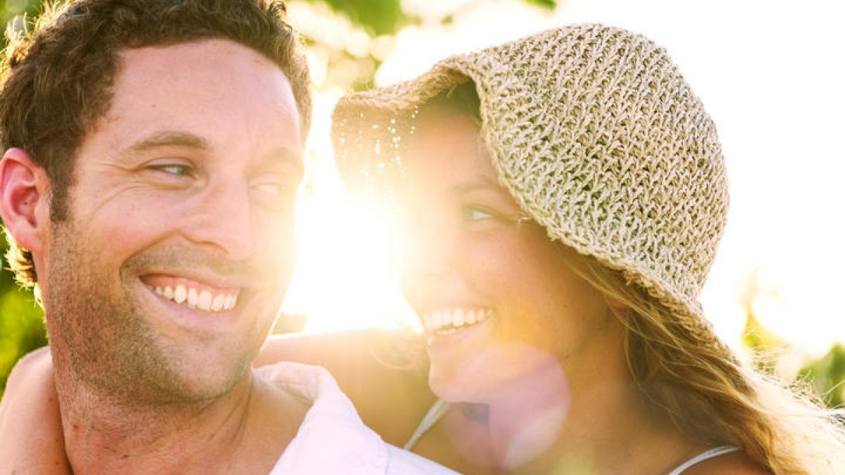 The image shows a couple smiling and looking at each other joyfully outdoors, with sunlight streaming from behind. Both appear happy and relaxed.