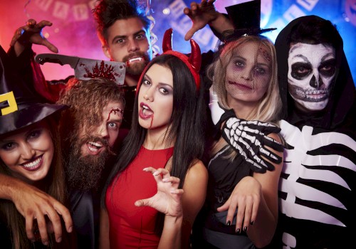 A group of people in Halloween costumes, including a witch, vampire, and skeleton, posing playfully with decor in the background.