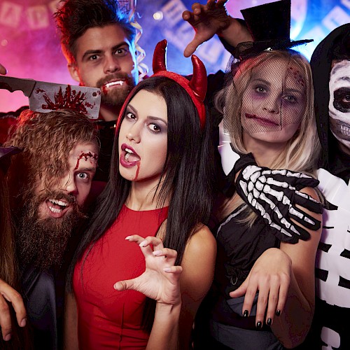 A group of people in Halloween costumes, including a witch, vampire, and skeleton, posing playfully with decor in the background.