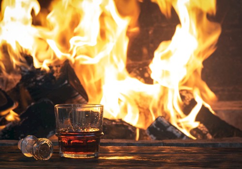 A glass of whiskey and a bottle stopper sit on a table in front of a roaring fireplace, creating a cozy ambiance.