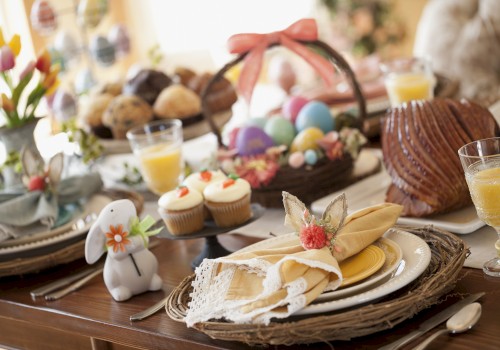 Festive table setting with Easter eggs, cupcakes, ham, and a ceramic bunny, surrounded by drinks and flowers.
