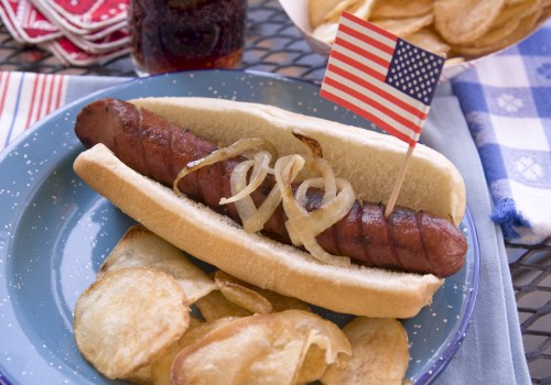 A grilled hot dog in a bun topped with onions and a small American flag, served with potato chips on a plate.