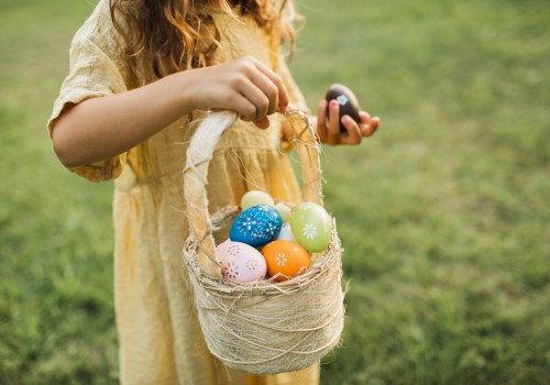 A child in a yellow dress holds a basket filled with colorful, decorated eggs, standing on grass.