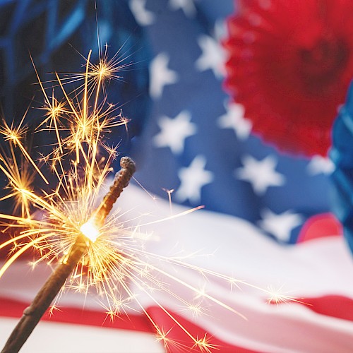 A lit sparkler in front of an American flag and festive decorations, creating a celebratory and patriotic scene.