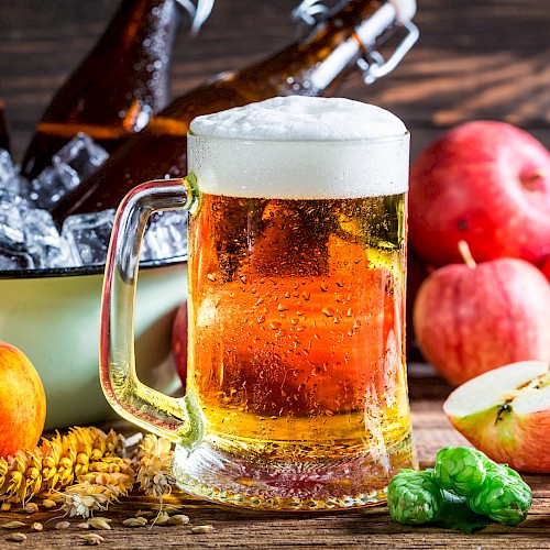 A glass of beer with a foamy head, bottles in ice, apples, wheat, and hops arranged on a wooden surface complete the scene.