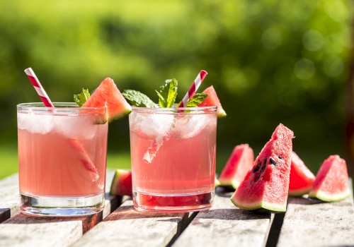 Two glasses of watermelon drinks with ice, garnished with watermelon slices and mint, placed on a wooden table outside.