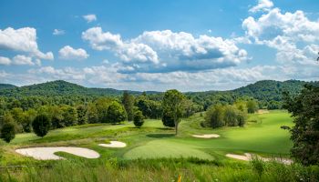 A scenic golf course with lush greenery, sand traps, and distant hills under a partly cloudy blue sky.