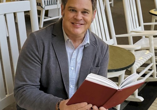 A person is sitting on a white rocking chair, holding an open book, in a room with more chairs and tables.
