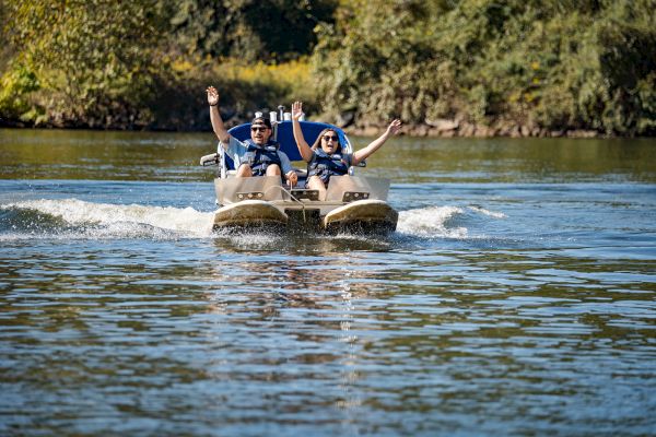 Two people are enjoying a ride on a pedal boat in a scenic lake, both raising their arms with joy. The background shows lush greenery.