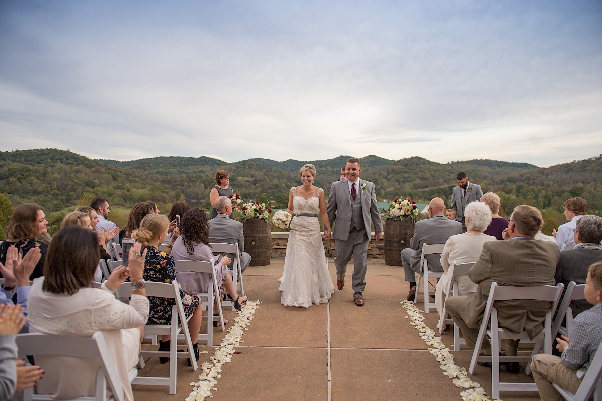 A couple walks down the aisle at an outdoor wedding ceremony with seated guests and scenic hills in the background.