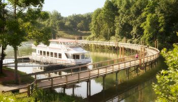 A white boat docks along a curved wooden boardwalk, surrounded by lush green trees and still waters reflecting the scenery.