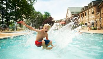 A boy in red and blue swim trunks is caught mid-air as he jumps into a swimming pool, creating a big splash, with buildings and people in the background.