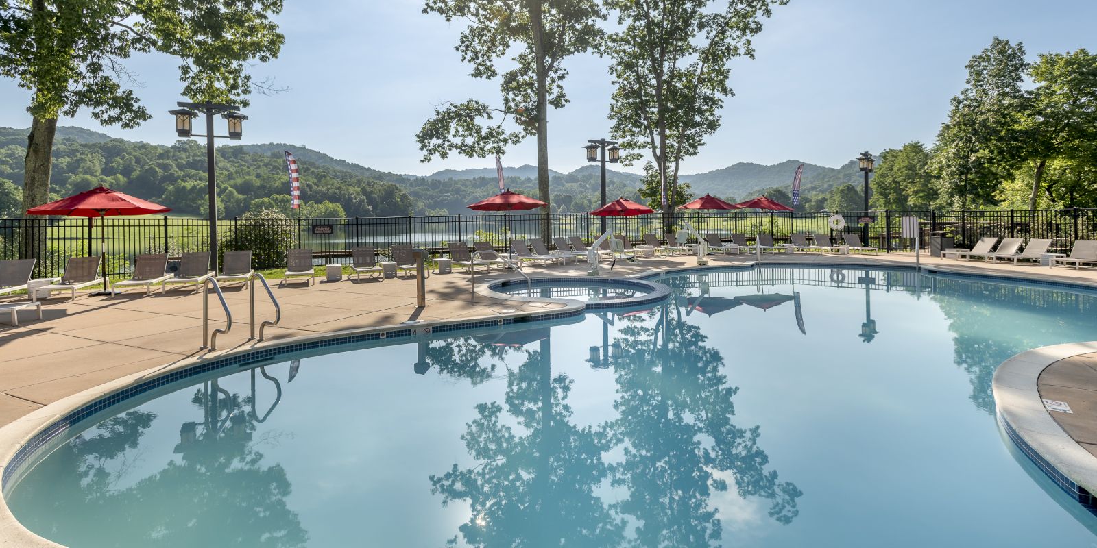 A beautiful outdoor swimming pool surrounded by lounge chairs and red umbrellas, with trees and mountains in the background.