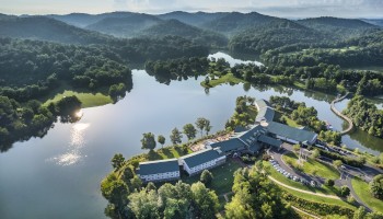Aerial view of a serene lakeside resort surrounded by lush green hills and forests, with buildings and roads nestled along the shoreline.