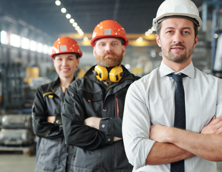 Three factory workers wearing safety equipment are standing in an industrial setting. The man in front is wearing a white hard hat and tie.