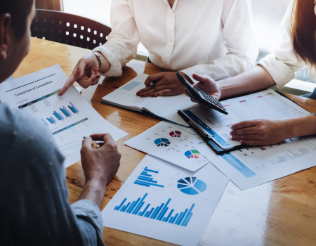 Three people are in a meeting, analyzing graphs and charts on paper documents and a laptop. One person is holding a calculator.